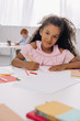 © LIGHTFIELD STUDIOS - selective focus of smiling african american kid at table with papers and pencils for drawing in classroom