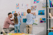 © LIGHTFIELD STUDIOS - smiling teacher helping little preschoolers hang colorful pictures on wall in classroom