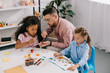 © LIGHTFIELD STUDIOS - teacher in eyeglasses and multiracial children drawing pictures with paints at table in classroom
