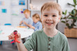 © LIGHTFIELD STUDIOS - selective focus of happy kid showing plasticine figure in hand with classmates behind in classroom