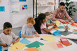 © LIGHTFIELD STUDIOS - teacher and multicultural preschoolers cutting colorful papers with scissors in classroom