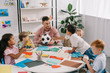 © LIGHTFIELD STUDIOS - male teacher with soccer ball and multiracial preschoolers sitting at table with colorful papers in classroom