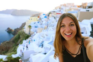 Wall Mural - Europe travel selfie smiling girl in Oia village, Santorini. Cute happy smiling tourist girl taking self-portrait picture during summer vacation in famous European destination Santorini, Greece.