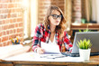 © proimagecontent - Happy attractive female architect in plaid shirt and glasses sitting and working with laptop in a loft office