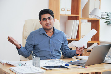 Confused Indian man sitting with hands apart and holding papers while looking at camera in office