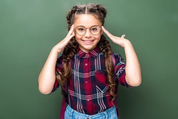 Wall Mural - smiling schoolchild touching head and looking at camera near blackboard