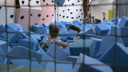 Wall Mural - Handheld shot of two multi ethnic girls jumping into cube pit in playroom and throwing blue soft foam cubes