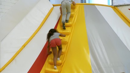 Wall Mural - Follow shot of young black girl ascending stairs of inflatable structure in indoor playground when spending time together with friends