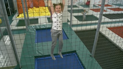 Wall Mural - Happy young girl jumping with hands in the air and bouncing on indoor trampoline when spending time with friends in play center, shot through net