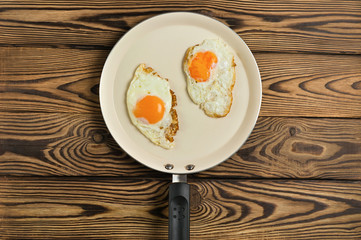 Two fried eggs in metal beige pan on old wooden brown table. Top view