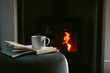 © Helen Rushbrook/Stocksy - Mug of hot chocolate and book in front of a lit stove