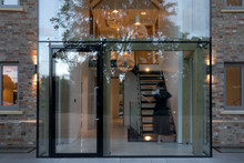 Evening View Of The Glass Entrance To A Modern House With Lights On And A Woman Ascending The Stairs.