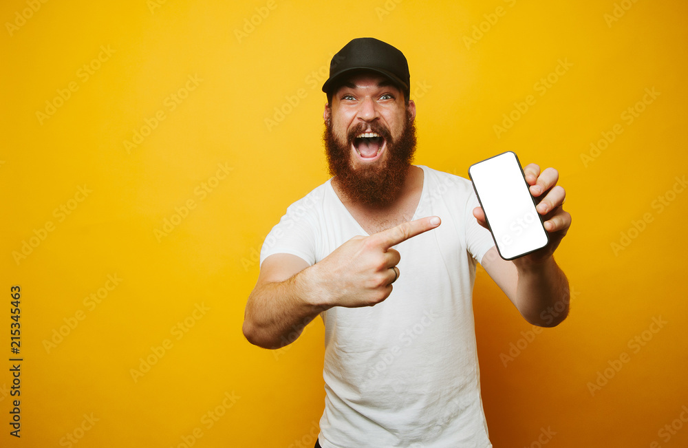 Amazed young bearded man in white t-shirt pointing at phone - obrazy, fototapety, plakaty 