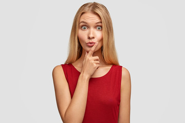 Pretty green eyed female stares with unexpected look, keeps mouth round, holds chin, looks surprisingly at something in front, expresses shock, wears casual red t shirt, isolated over white background