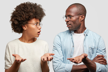 Wall Mural - Confused African American couple look at each other angrily, clasp hands, don`t know where their money disappeared, wear round spectacles, isolated over white background. Family has problems