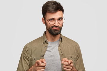 Wall Mural - Confident bearded man points at camera with both index fingers, selects something, has dark stubble, stands alone against white background. Attractive self assured ushaven male expresses his choice