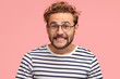 © WHstudio Leushin N - Joyful Caucasian male clenches teeth and looks positively at camera, has curly hair, wears spectacles and striped sweater, isolated over pink background. Happy man freelancer rejoices success