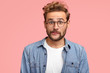 © wayhome.studio - Thrilled curly hipster with puzzled expression purses lips and looks with stunned expression, wears denim shirt, has stubble, isolated over pink background. People, reaction and lifestyle concept