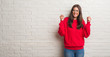 © Krakenimages.com - Young brunette woman standing over white brick wall excited for success with arms raised celebrating victory smiling. Winner concept.