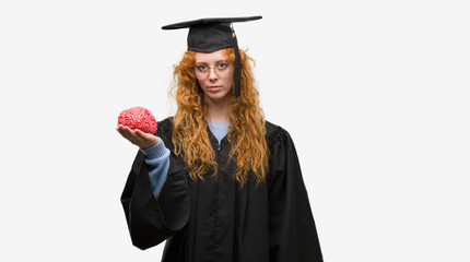 Sticker - Young redhead student woman wearing graduated uniform holding brain with a confident expression on smart face thinking serious