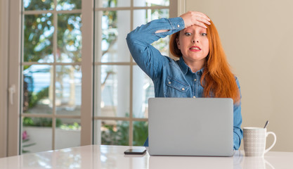 Redhead woman using computer laptop at home stressed with hand on head, shocked with shame and surprise face, angry and frustrated. Fear and upset for mistake.