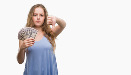 Canvas Print - Young blonde woman holding dollars with angry face, negative sign showing dislike with thumbs down, rejection concept