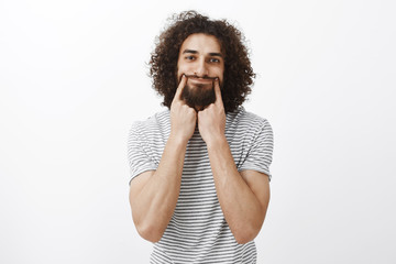 Wall Mural - Portrait of depressed unhappy handsome hispanic bearded guy with curly hair, pulling smile with index fingers, trying look positive while feeling upset and sad, standing intense over gray background