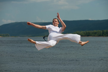 Young guy trains capoeira on sky backround. A men performs martial the kick in the jump