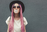Fototapeta  - Stylish young hipster woman with long pink hair blowing a bubble with bubble gum.