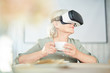 © pressmaster - Modern senior female with virtual reality headset sitting by table and having tea or coffee
