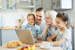 © pressmaster - Happy young family of four sitting in front of laptop and talking through video-chat in the kitchen