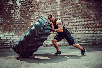 Muscular bearded tattooed fitness man moving large tire in street gym. Concept lifting, workout training.