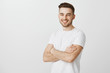 Portrait of pleased confident and accomplished european guy with stylish haircut standing unshaven over gray background crossing hands on chest smiling joyfully and satisfied at camera