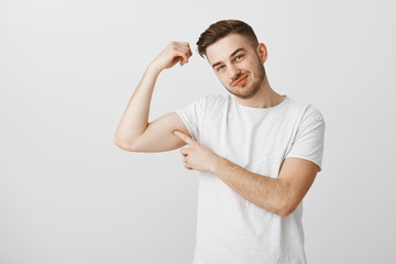 Wall Mural - Wanna touch my strong biceps. Cute and flirty handsome male with beard in white casual t-shirt tilting head and smiling with delighted and pleased look poking himself in mascule on right hand