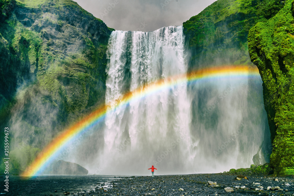Iceland waterfall travel nature famous tourist destination. Skogafoss waterfall with rainbow and woman under water fall in magical landscape popular Europe attraction. - obrazy, fototapety, plakaty 