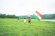 3 Cute Little Indian Kids Holding, Waving Or Running With Tricolour Near A Lake With Greenery In The Background, Celebrating Independence Or Republic Day  