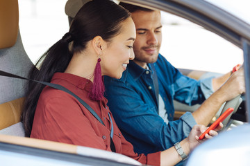 Wall Mural - Modern generation. Delighted Asian woman using her smartphone while sitting in the car with her boyfriend