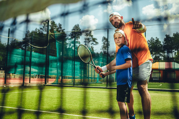 Portrait of concentrated male trainer teaching serene boy playing tennis outdoor. He pointing hand