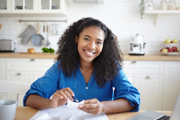 People, paperwork and modern technology concept. Picture of joyful young African woman holding pen and bill while calculating family budget at home, sitting in kitchen with papers and laptop on table