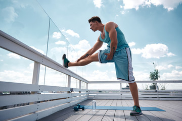 Wall Mural - Fit young man is stretching leg while standing and putting it on roof railing. He is getting ready for training outdoors with dumbbells. Male is working out in fresh air concept