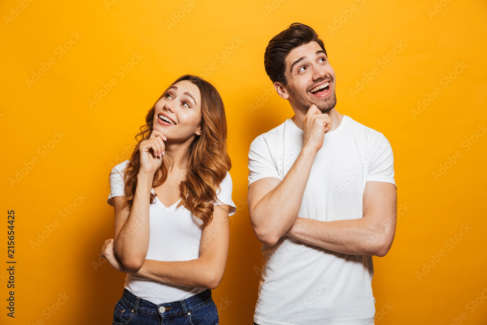 Image of cheerful young people man and woman in basic clothing laughing and touching chin while looking aside, isolated over yellow background - obrazy, fototapety, plakaty 