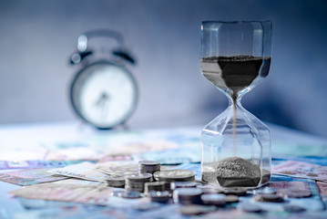 Sand running through the shape of hourglass on table with banknotes and coins of international currency. Time investment and retirement saving. Urgency countdown timer for business deadline concept