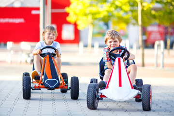 Two active little kid boys driving pedal race car in summer garden, outdoors. Children, best friends racing with fast speed and having fun.