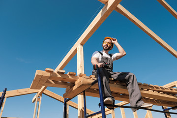 The man builder sits on the edge of the roof of the frame house, in a yellow helmet and gray overalls. The blue sky and clear sunny day.