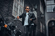 © Fxquadro - Fashionable biker in sunglasses dressed in a black leather jacket and jeans leaning on his retro motorcycle on an old Europe street.