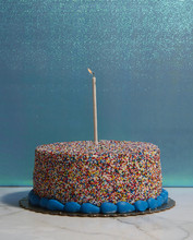 Sprinkled Round Cake With One Burning Candle On Blue Background.
