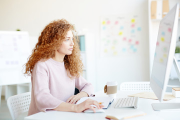 Serious confident curly-haired female designer in casual sweater sitting at table and using desktop computer in modern office