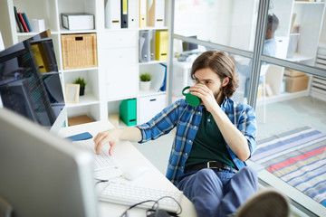 Wall Mural - Serious relaxed hipster young software engineer in casual clothing working in office: he keeping feet on table and drinking coffee while installing program on computer