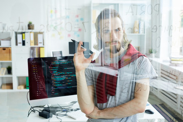 Wall Mural - Serious confident handsome young computer programmer with beard developing code for software and holding felt tip pen while looking at data in glassy board in office.