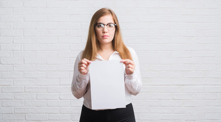 Wall Mural - Young adult business woman standing over white brick wall holding blank paper with a confident expression on smart face thinking serious